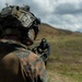 A military service member in full combat gear is seen from behind, aiming a rifle with an optical sight across an open, hilly terrain. The individual is wearing MARPAT (Marine pattern) camouflage uniform and a tactical helmet equipped with night vision device mounts. The setting appears to be an outdoor training range or field exercise environment.