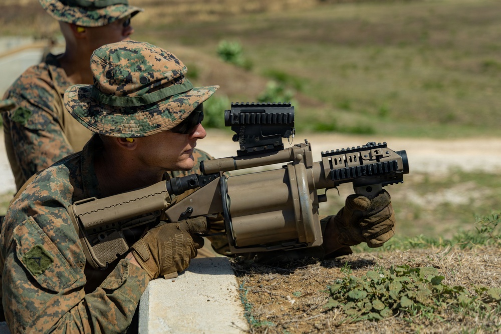 A U.S. Marine in MARPAT digital camouflage uniform is aiming a multi-shot grenade launcher, likely an M32 MGL (Multiple Grenade Launcher), at a firing range. The weapon features a tan/desert finish with a drum-style cylinder and mounted optics. A second Marine in similar uniform is visible in the background.