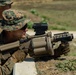 A U.S. Marine in MARPAT digital camouflage uniform is aiming a multi-shot grenade launcher, likely an M32 MGL (Multiple Grenade Launcher), at a firing range. The weapon features a tan/desert finish with a drum-style cylinder and mounted optics. A second Marine in similar uniform is visible in the background.