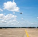 A military airfield tarmac scene showing a helicopter in flight above a runway marked with a yellow centerline. Two red military fire/crash trucks are positioned on either side of the tarmac, with additional helicopters and aircraft visible in the background. The setting appears to be a U.S. Army installation, likely Fort Stewart based on visible markings on the fire trucks.