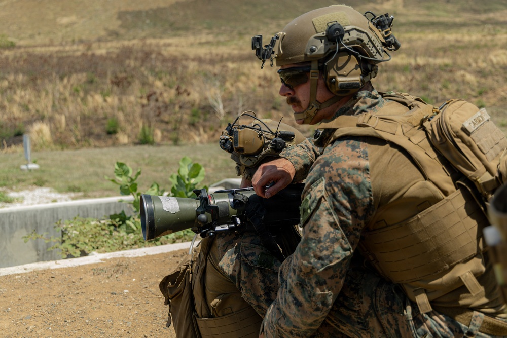 Two military personnel in MARPAT (Marine pattern) camouflage uniforms are positioned in a crouching stance, with one operating what appears to be a shoulder-fired rocket launcher or recoilless weapon system. Both individuals are wearing tactical helmets with communications equipment, ballistic eyewear, and full combat gear including plate carriers. The setting appears to be an outdoor training range or operational environment with arid terrain in the background.