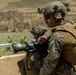 Two military personnel in MARPAT (Marine pattern) camouflage uniforms are positioned in a crouching stance, with one operating what appears to be a shoulder-fired rocket launcher or recoilless weapon system. Both individuals are wearing tactical helmets with communications equipment, ballistic eyewear, and full combat gear including plate carriers. The setting appears to be an outdoor training range or operational environment with arid terrain in the background.