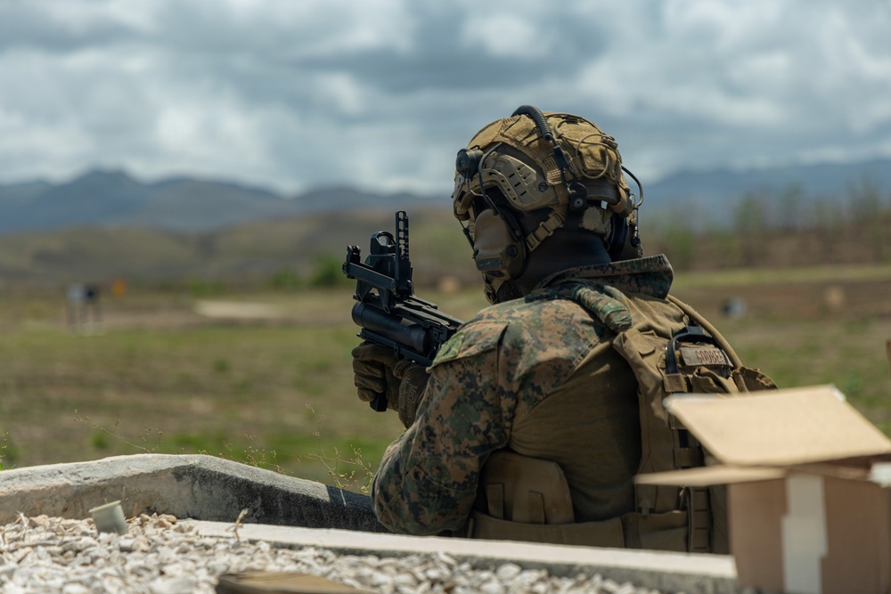 A military service member in full combat gear is positioned behind a concrete barrier at what appears to be a live-fire training range. The individual is aiming a rifle downrange toward distant targets visible in the background. The setting appears to be an outdoor training facility with mountainous terrain in the distance.