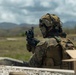 A military service member in full combat gear is positioned behind a concrete barrier at what appears to be a live-fire training range. The individual is aiming a rifle downrange toward distant targets visible in the background. The setting appears to be an outdoor training facility with mountainous terrain in the distance.