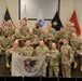 A group of approximately 18 U.S. Army soldiers in OCP (Operational Camouflage Pattern) uniforms pose for a formal group photograph. Several soldiers in the front row are holding an Illinois state flag. The setting appears to be an indoor military facility or conference room with multiple flags displayed in the background.