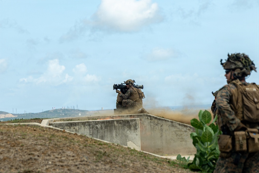 Two military personnel in full combat gear are positioned behind a concrete barrier in what appears to be a live-fire training exercise. One soldier is actively firing a shoulder-mounted rocket or missile launcher, evidenced by the visible backblast dust cloud and smoke trail above. The setting appears to be a military training range with hilly terrain visible in the background.