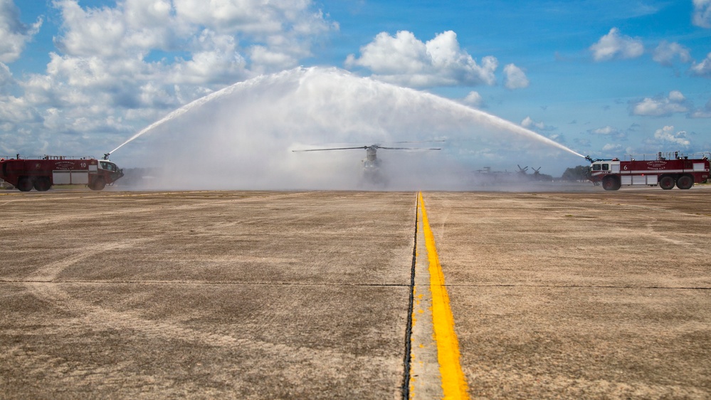 A military water salute ceremony is taking place on an airfield tarmac, with two red fire/crash trucks spraying arching water streams over a large military helicopter (appears to be a CH-47 Chinook or similar tandem-rotor aircraft). The yellow centerline of the taxiway leads directly toward the aircraft positioned between the two trucks. This type of ceremony typically honors a final flight or retirement of an aircraft or crew member.