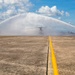 A military water salute ceremony is taking place on an airfield tarmac, with two red fire/crash trucks spraying arching water streams over a large military helicopter (appears to be a CH-47 Chinook or similar tandem-rotor aircraft). The yellow centerline of the taxiway leads directly toward the aircraft positioned between the two trucks. This type of ceremony typically honors a final flight or retirement of an aircraft or crew member.