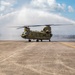 A military CH-47 Chinook tandem-rotor helicopter is receiving a traditional water cannon salute on a tarmac, likely marking a retirement, final flight, or ceremonial occasion. Two red fire/crash trucks (one visibly numbered '10') flank the aircraft on either side, projecting arching water streams that form a ceremonial arch over the helicopter. The scene takes place on a concrete airfield apron under a partly cloudy sky.