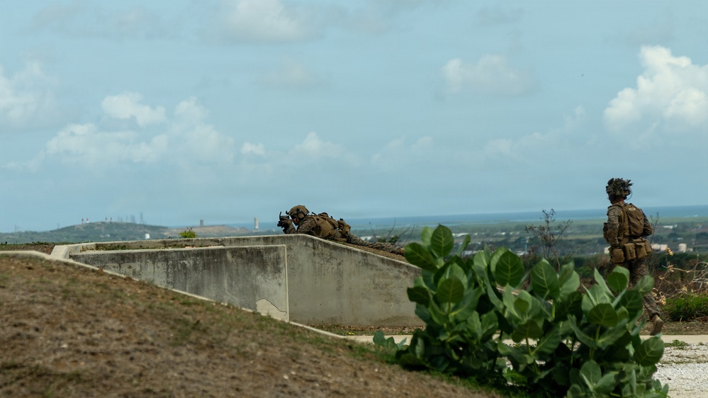 The image shows military personnel in full combat gear conducting what appears to be a tactical training exercise on elevated terrain. One soldier is in a prone position behind a concrete wall structure, aiming downrange, while a second soldier stands nearby in an overwatch position. The background reveals a coastal or lowland area with industrial structures visible on the horizon.