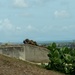 The image shows military personnel in full combat gear conducting what appears to be a tactical training exercise on elevated terrain. One soldier is in a prone position behind a concrete wall structure, aiming downrange, while a second soldier stands nearby in an overwatch position. The background reveals a coastal or lowland area with industrial structures visible on the horizon.