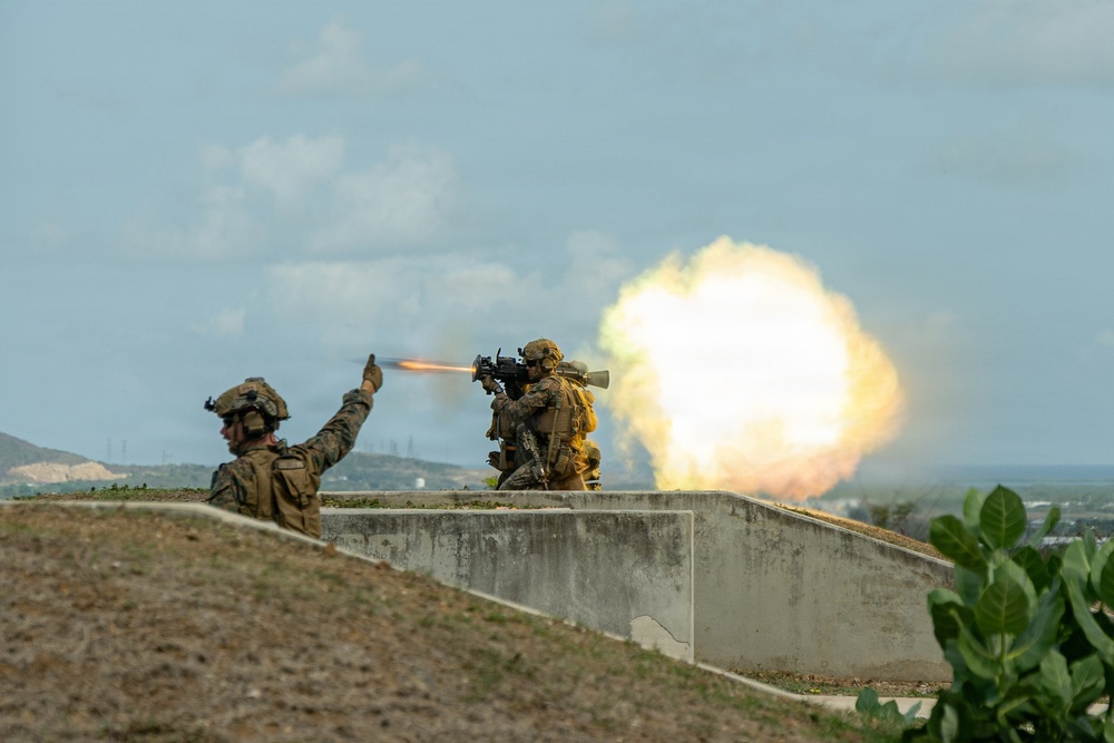Two soldiers in full combat gear are positioned behind a concrete barrier, with one firing a shoulder-launched rocket or missile system. A large fireball and exhaust plume are visible to the right of the shooter, indicating the weapon has just been discharged. The second soldier has his arm raised, likely performing a safety backblast area clearance signal.