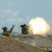 Two soldiers in full combat gear are positioned behind a concrete barrier, with one firing a shoulder-launched rocket or missile system. A large fireball and exhaust plume are visible to the right of the shooter, indicating the weapon has just been discharged. The second soldier has his arm raised, likely performing a safety backblast area clearance signal.
