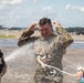 A military service member in camouflage uniform is being doused with water on what appears to be an airfield tarmac. Two civilians in colorful clothing are visible in the foreground, one holding a hose spraying water. This appears to be a traditional military 'fini flight' or homecoming water salute ceremony.