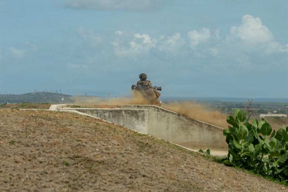 A military personnel is positioned behind a concrete barrier or berm, firing what appears to be a shoulder-launched weapon system, generating a large cloud of dust and backblast. The scene appears to be a training range or firing position, with an open arid landscape and distant hills visible in the background. Green tropical vegetation is visible in the foreground right.