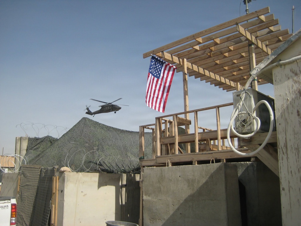 The image shows a forward operating base or military installation with an American flag hanging vertically from a wooden structure under construction. A military helicopter, consistent with a UH-60 Black Hawk, is visible in flight in the background. The scene includes typical base security features such as concertina wire and concrete barriers (T-walls/HESCO barriers).