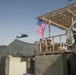 The image shows a forward operating base or military installation with an American flag hanging vertically from a wooden structure under construction. A military helicopter, consistent with a UH-60 Black Hawk, is visible in flight in the background. The scene includes typical base security features such as concertina wire and concrete barriers (T-walls/HESCO barriers).