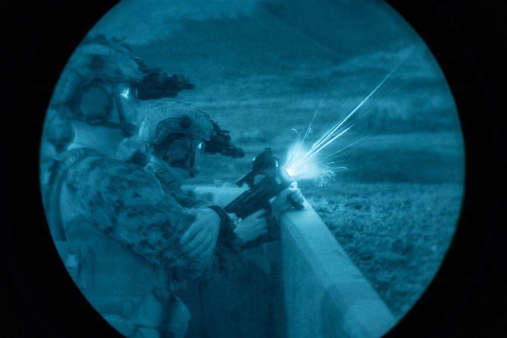 A night-vision scope image showing at least two military personnel in full combat gear firing a weapon over a barrier or wall. A visible muzzle flash is captured at the moment of discharge, illuminating the scene. The circular vignette and blue-green tonal rendering are characteristic of night-vision optics.