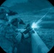 A night-vision scope image showing at least two military personnel in full combat gear firing a weapon over a barrier or wall. A visible muzzle flash is captured at the moment of discharge, illuminating the scene. The circular vignette and blue-green tonal rendering are characteristic of night-vision optics.