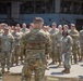 A military formation or gathering is taking place outdoors in front of a large industrial hangar or warehouse structure. One soldier, seen from behind, appears to be addressing a large group of uniformed personnel standing in a semi-circular arrangement. The setting and attire suggest a formal military ceremony or briefing event.