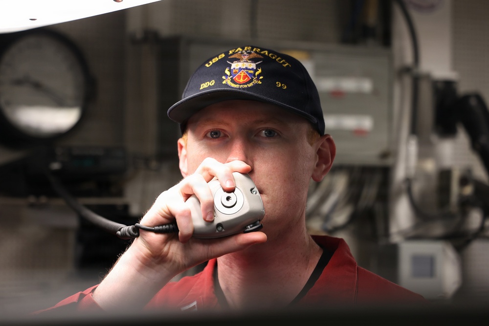 A U.S. Navy sailor aboard USS Farragut (DDG-99) is using a shipboard communication handset, likely a 1MC general announcing system or sound-powered phone. He is wearing a navy blue ball cap embroidered with 'USS FARRAGUT DDG 99' and a red uniform shirt. The background shows shipboard equipment consistent with an interior naval vessel compartment.