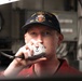 A U.S. Navy sailor aboard USS Farragut (DDG-99) is using a shipboard communication handset, likely a 1MC general announcing system or sound-powered phone. He is wearing a navy blue ball cap embroidered with 'USS FARRAGUT DDG 99' and a red uniform shirt. The background shows shipboard equipment consistent with an interior naval vessel compartment.