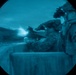 The image is captured through a night vision or thermal optic lens, showing a circular field of view with a blue-green tint characteristic of image intensification technology. Two military personnel in camouflage uniforms are visible, with one aiming a weapon equipped with a tactical light or laser that is actively emitting a bright beam. Both individuals are wearing night vision devices mounted on their helmets.