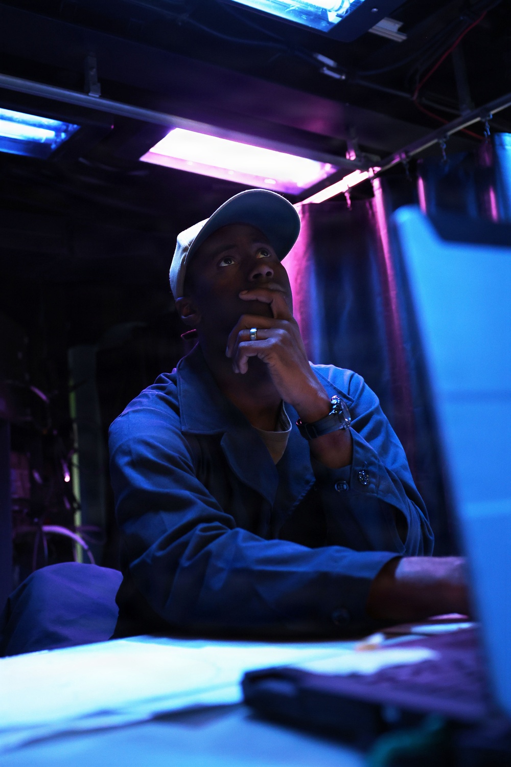 A man sits at a desk or workstation in a dimly lit environment illuminated by blue and purple/pink neon or fluorescent lighting. He is wearing a blue jacket and a cap, with his hand raised to his chin in a contemplative or thoughtful pose. The setting appears to be a backstage, studio, or technical workspace.