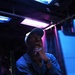 A man sits at a desk or workstation in a dimly lit environment illuminated by blue and purple/pink neon or fluorescent lighting. He is wearing a blue jacket and a cap, with his hand raised to his chin in a contemplative or thoughtful pose. The setting appears to be a backstage, studio, or technical workspace.