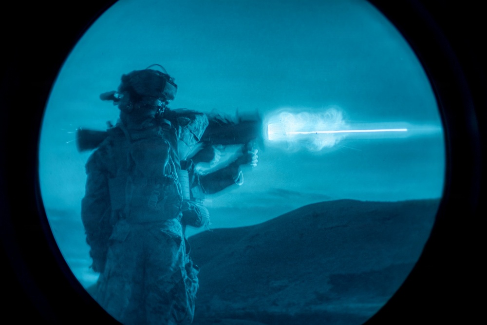 A soldier in full combat gear is firing a weapon, captured through what appears to be a night-vision or thermal imaging device, giving the image a distinctive blue monochromatic appearance. A bright muzzle flash and smoke are clearly visible at the barrel of the weapon. The background shows a desert or arid terrain landscape at night.