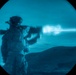 A soldier in full combat gear is firing a weapon, captured through what appears to be a night-vision or thermal imaging device, giving the image a distinctive blue monochromatic appearance. A bright muzzle flash and smoke are clearly visible at the barrel of the weapon. The background shows a desert or arid terrain landscape at night.