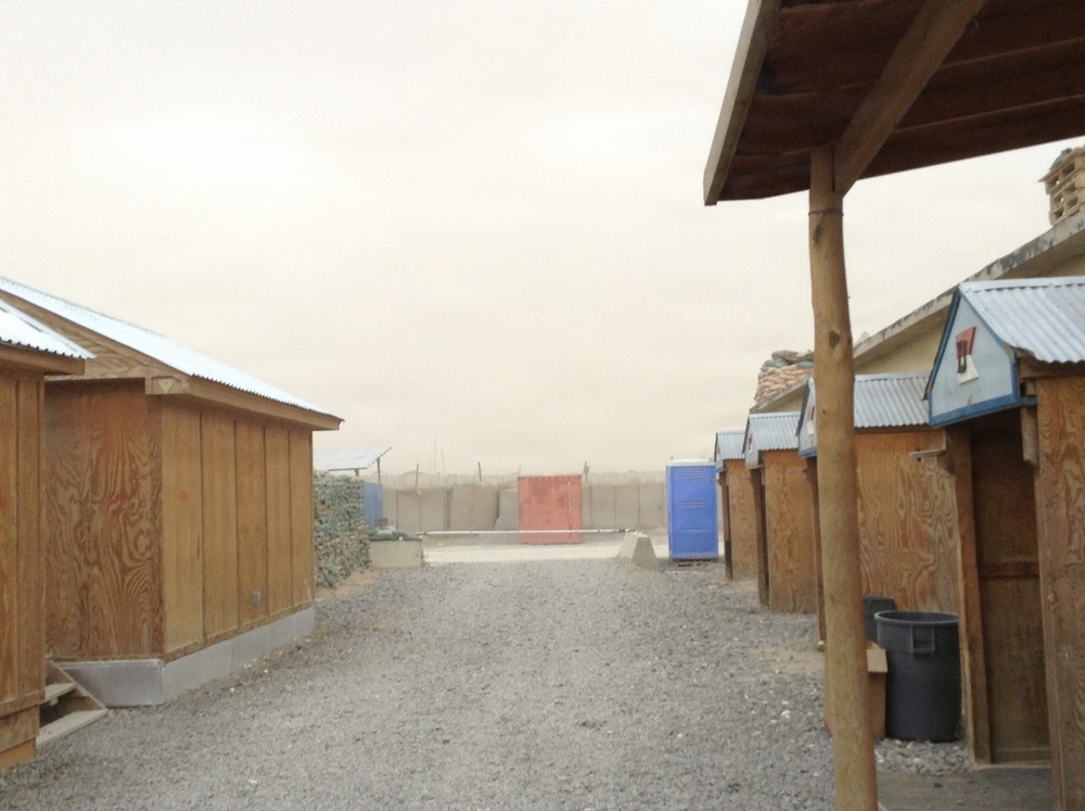 The image shows a forward operating base or military camp compound during a dust storm or sandstorm, with visibility significantly reduced by airborne particulate matter. Rows of plywood huts with corrugated metal roofs line a gravel pathway, with portable toilets visible in the background. The overall atmosphere suggests an arid, desert environment consistent with military installations in regions such as Afghanistan or Iraq.