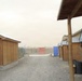 The image shows a forward operating base or military camp compound during a dust storm or sandstorm, with visibility significantly reduced by airborne particulate matter. Rows of plywood huts with corrugated metal roofs line a gravel pathway, with portable toilets visible in the background. The overall atmosphere suggests an arid, desert environment consistent with military installations in regions such as Afghanistan or Iraq.