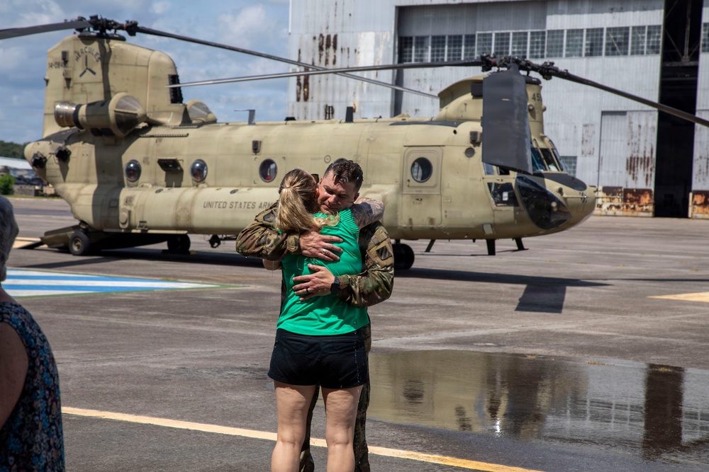 A United States Army soldier in camouflage uniform embraces a woman in a green shirt on a wet tarmac in front of a CH-47 Chinook helicopter. The scene appears to be a military homecoming or reunion, taking place at what looks like a military airfield or hangar facility. The emotional embrace and the presence of the large transport helicopter suggest the soldier has recently returned from a deployment.