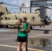 A United States Army soldier in camouflage uniform embraces a woman in a green shirt on a wet tarmac in front of a CH-47 Chinook helicopter. The scene appears to be a military homecoming or reunion, taking place at what looks like a military airfield or hangar facility. The emotional embrace and the presence of the large transport helicopter suggest the soldier has recently returned from a deployment.