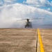 A military tandem-rotor helicopter, consistent with a CH-47 Chinook, is on a tarmac or airfield surface being subjected to a water salute or wash-down arc. The rotor wash is dispersing the water spray into a dramatic mist cloud surrounding the aircraft. A yellow centerline stripe is visible in the foreground of the concrete surface.