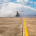 A military tandem-rotor helicopter, consistent with a CH-47 Chinook, is receiving a ceremonial water salute on a military airfield tarmac. Two fire trucks are positioned on either side of the yellow centerline, projecting arching water streams that form a tunnel over the helicopter. This type of water arch ceremony typically marks a significant milestone such as a final flight or retirement of an aircraft.