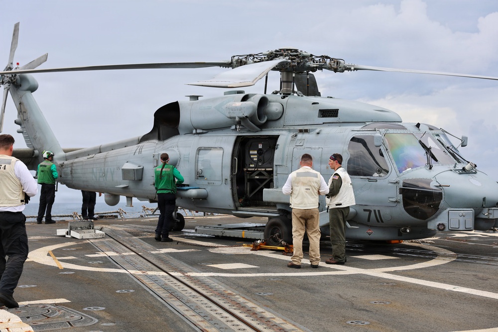 A U.S. Navy helicopter, marked with the number '711' and 'NAVY' on its fuselage, is parked on the flight deck of a naval vessel. Several personnel in various colored vests and uniforms are gathered around the aircraft, apparently conducting inspection or maintenance activities. The helicopter appears to be an MH-60 Seahawk variant with its side door open.
