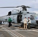 A U.S. Navy helicopter, marked with the number '711' and 'NAVY' on its fuselage, is parked on the flight deck of a naval vessel. Several personnel in various colored vests and uniforms are gathered around the aircraft, apparently conducting inspection or maintenance activities. The helicopter appears to be an MH-60 Seahawk variant with its side door open.