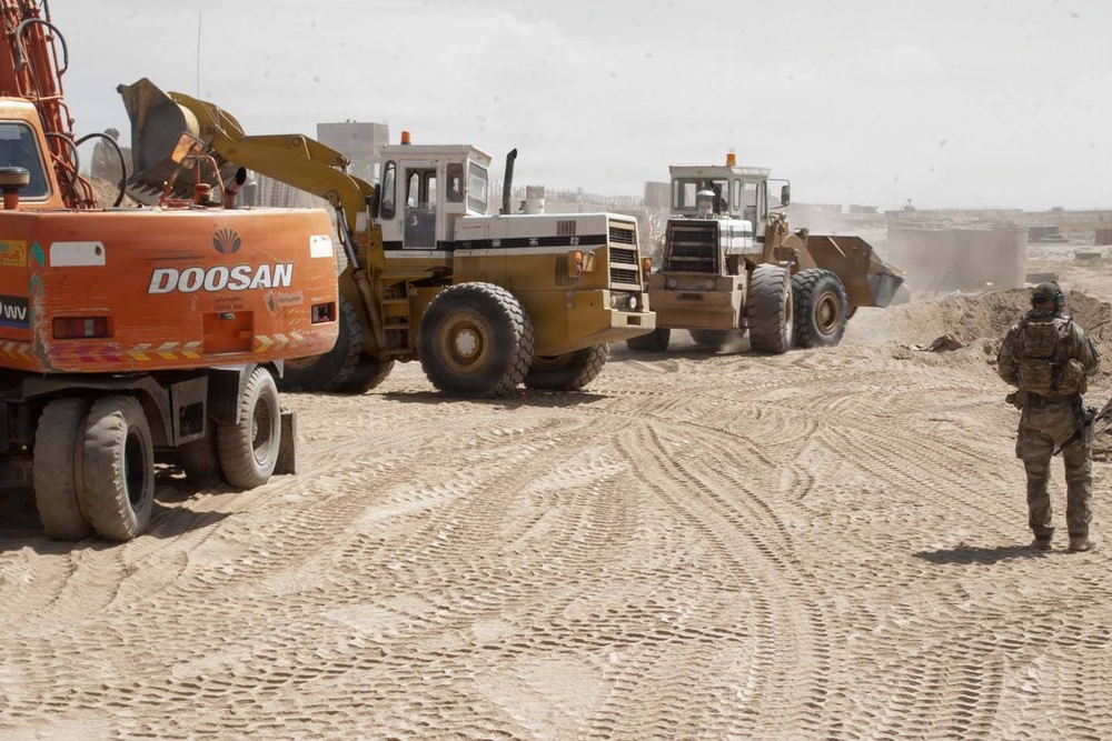 A military soldier in full combat gear observes construction equipment operating on a dusty, arid terrain. Three pieces of heavy machinery are visible, including a Doosan excavator and two wheel loaders actively moving earth. The setting appears to be a construction or infrastructure project in a desert environment, likely in the Middle East.