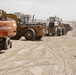 A military soldier in full combat gear observes construction equipment operating on a dusty, arid terrain. Three pieces of heavy machinery are visible, including a Doosan excavator and two wheel loaders actively moving earth. The setting appears to be a construction or infrastructure project in a desert environment, likely in the Middle East.