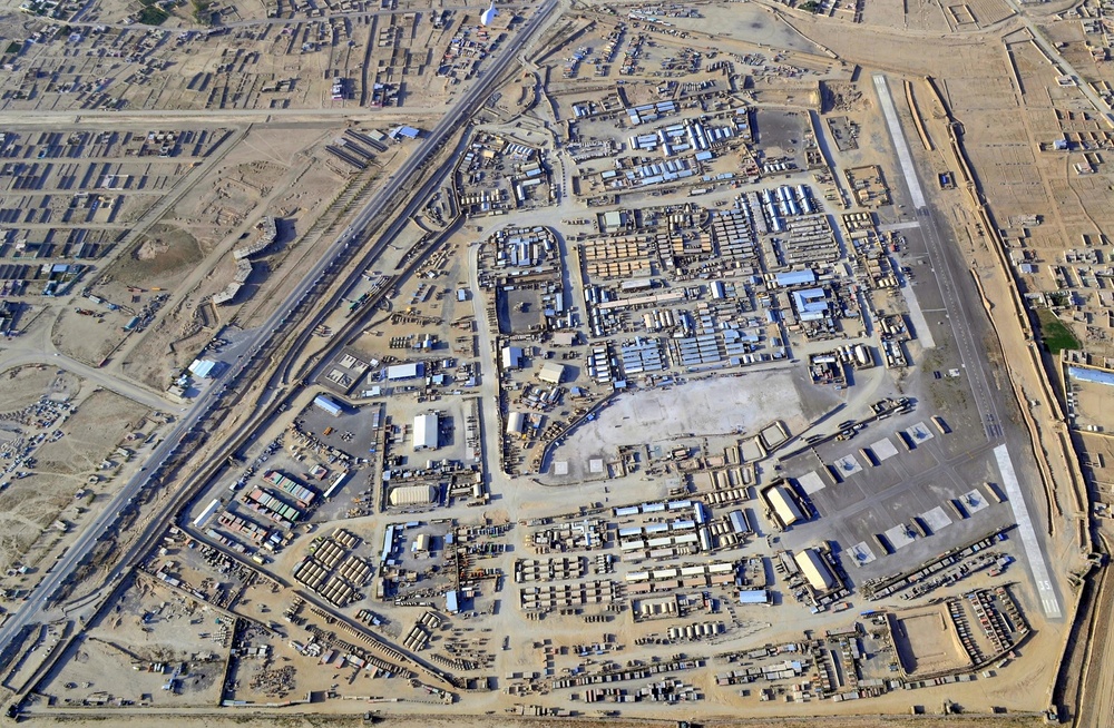 Aerial photograph of a large military forward operating base (FOB) or logistics installation situated adjacent to an urban area in an arid environment. The compound contains numerous modular/prefabricated structures, storage areas, and what appears to be a runway or taxiway along the eastern perimeter. The surrounding area shows dense low-rise urban development typical of Central or Southwest Asian cities.