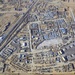 Aerial photograph of a large military forward operating base (FOB) or logistics installation situated adjacent to an urban area in an arid environment. The compound contains numerous modular/prefabricated structures, storage areas, and what appears to be a runway or taxiway along the eastern perimeter. The surrounding area shows dense low-rise urban development typical of Central or Southwest Asian cities.