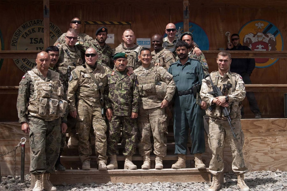 A group of approximately 13 military personnel pose for a formal group photograph in front of a wooden structure bearing the text 'Polish Military Contingent Task Force ISAF'. The group includes personnel in various camouflage uniforms and one individual in civilian-style clothing, suggesting a multinational military gathering. The setting appears to be a forward operating base or military compound.