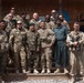 A group of approximately 13 military personnel pose for a formal group photograph in front of a wooden structure bearing the text 'Polish Military Contingent Task Force ISAF'. The group includes personnel in various camouflage uniforms and one individual in civilian-style clothing, suggesting a multinational military gathering. The setting appears to be a forward operating base or military compound.