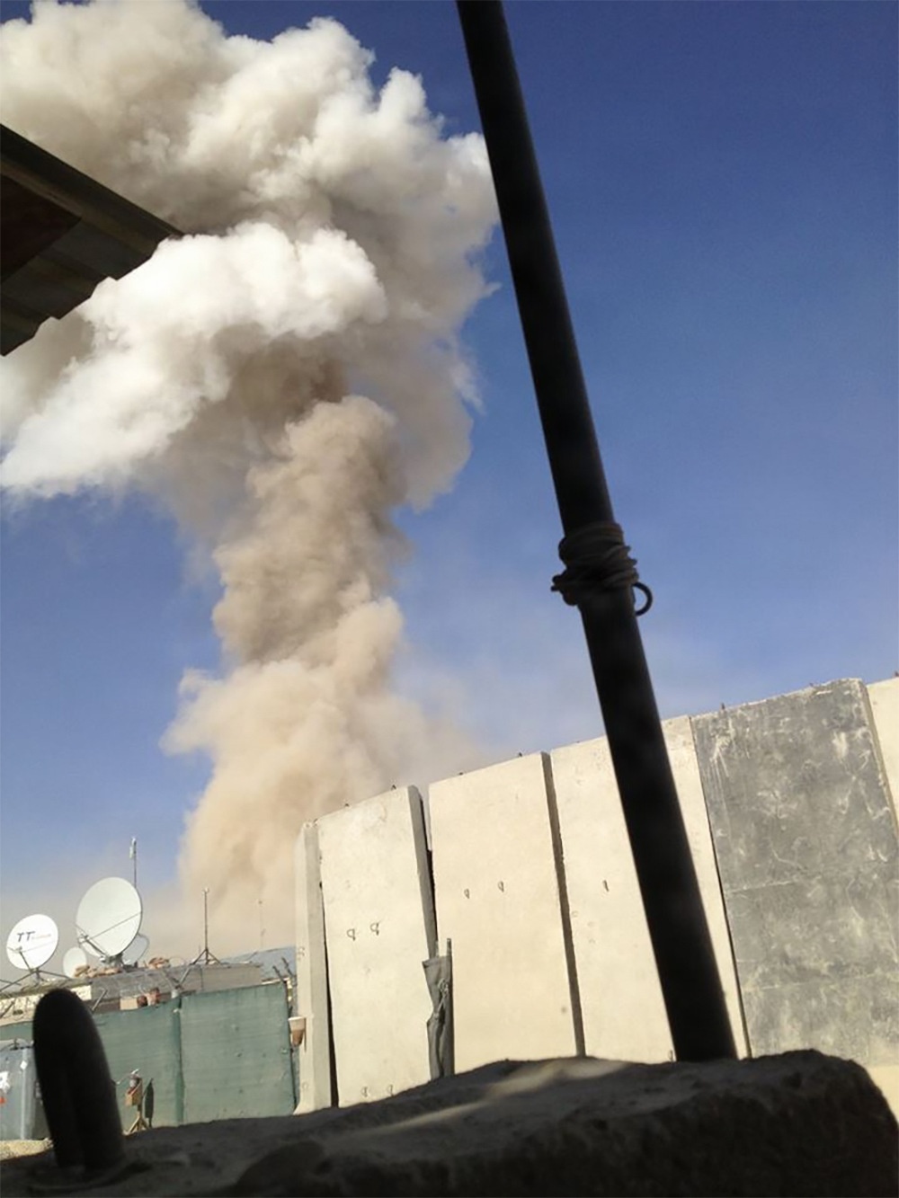 The image shows a large plume of smoke and dust rising into a clear blue sky, photographed from behind a fortified position. Concrete blast walls (T-walls) are visible in the foreground, along with satellite dishes and low buildings in the background. The scene suggests an explosion or detonation has occurred nearby.