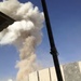 The image shows a large plume of smoke and dust rising into a clear blue sky, photographed from behind a fortified position. Concrete blast walls (T-walls) are visible in the foreground, along with satellite dishes and low buildings in the background. The scene suggests an explosion or detonation has occurred nearby.