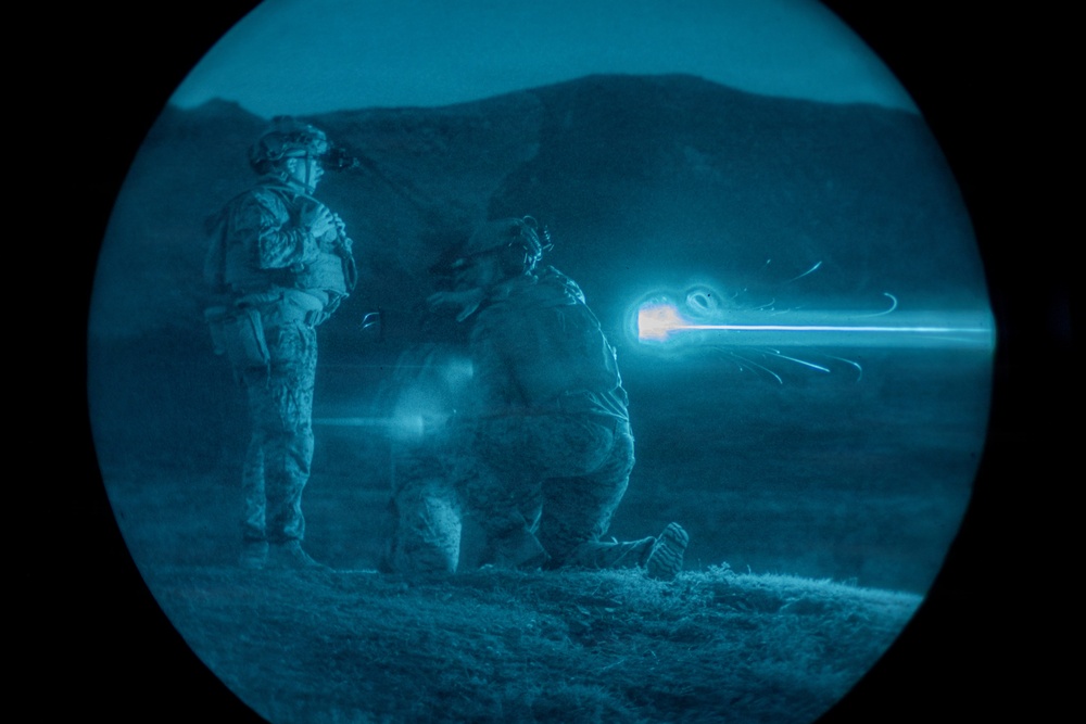 A night-vision captured image showing two military personnel in full combat gear in an outdoor terrain setting. One soldier is standing while another is in a kneeling firing position, with a visible muzzle flash and tracer round captured mid-flight. The circular vignette is characteristic of night-vision optic photography.