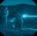 A night-vision captured image showing two military personnel in full combat gear in an outdoor terrain setting. One soldier is standing while another is in a kneeling firing position, with a visible muzzle flash and tracer round captured mid-flight. The circular vignette is characteristic of night-vision optic photography.