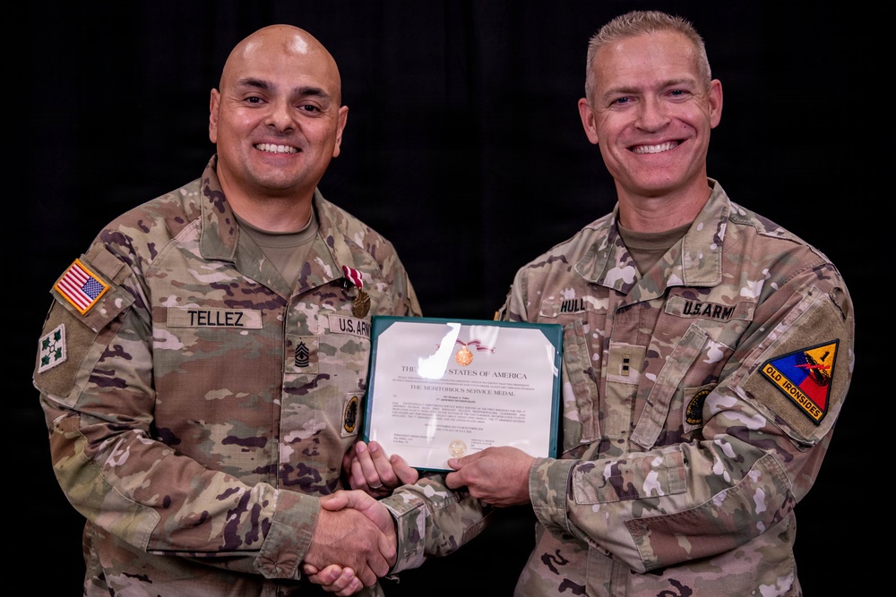 Two U.S. Army soldiers pose together holding a Meritorious Service Medal certificate. The soldier on the left, identified by name tape as 'TELLEZ,' is receiving the award and is wearing the medal on his uniform. The soldier on the right, identified as 'HULL,' appears to be the presenting officer, and both are shaking hands for the ceremonial photo.